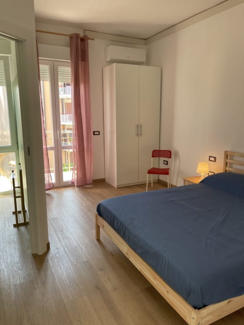 Camera luminosa degli Aparthotel Rigiariloo con letto in legno, armadio bianco e vista sulla città attraverso tende rosa a San