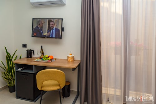 Scrivania d'hotel con sedia gialla, TV a parete, frutta fresca e tende con vista città nelle camere Macramè Fazello, Sciacca
