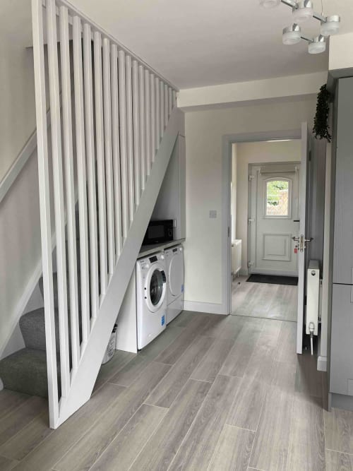 Modern laundry room at Duffy Lodge with white washing machine under stairs, grey tile flooring, white railing, and entrance