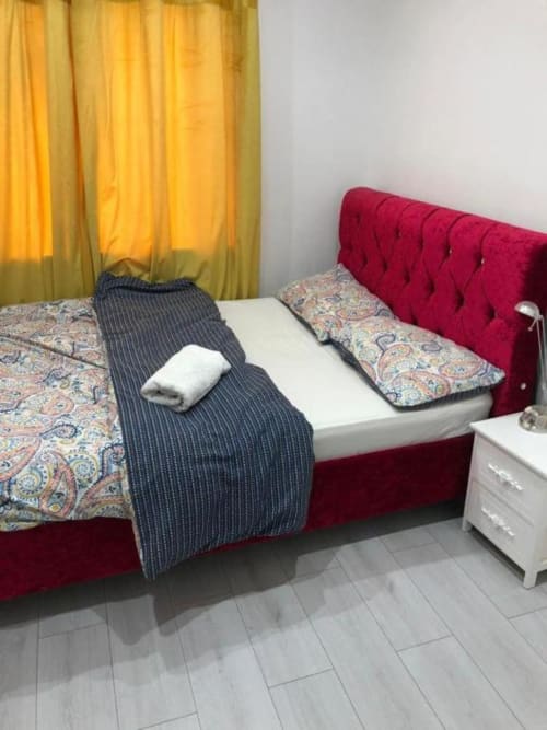 Bedroom with red upholstered bed, patterned bedding, white pillow, dark blanket, yellow curtains, white nightstand, and tile