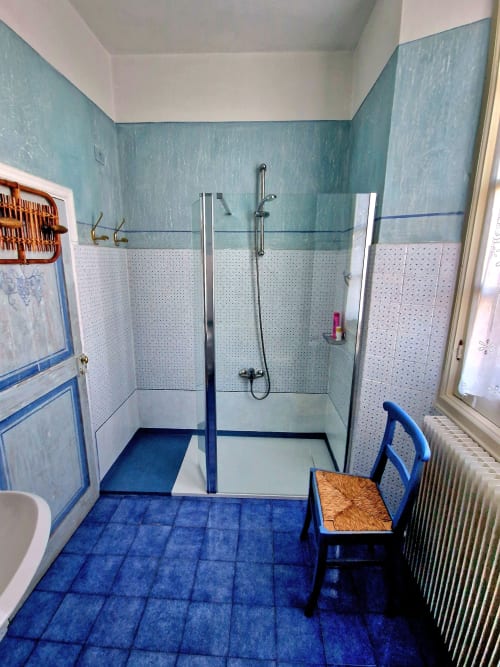 Bagno del B&B La Corte Ovada con pavimento in piastrelle blu, pareti turchesi, box doccia in vetro e sedia rustica.
