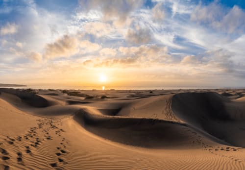 Puesta de sol sobre las dunas de arena de Maspalomas con huellas en la arena dorada del desierto y el horizonte del océano