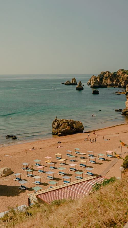 Praia dourada com espreguiçadeiras cobertas, formações rochosas e águas turquesas do albergue Ocean View Lagos em Portugal