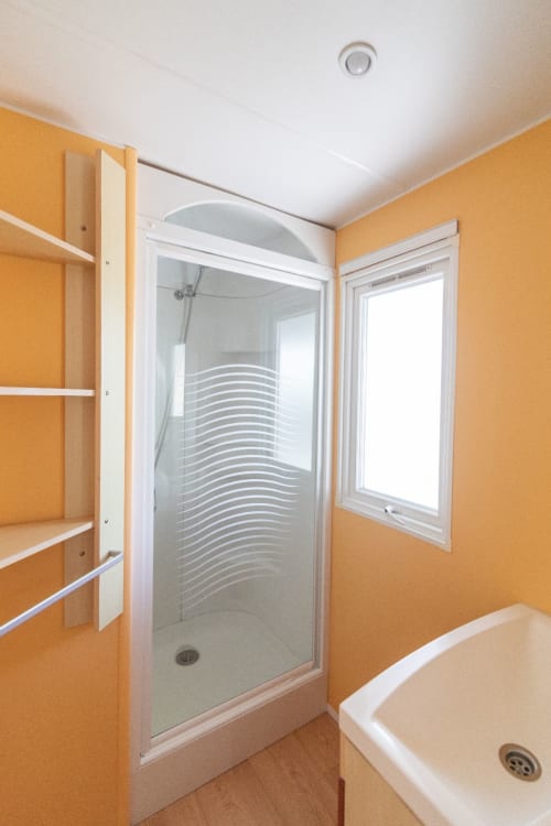 Modern bathroom with orange walls, glass shower enclosure, white fixtures, and window at Bungalow Buda Beach, Chipiona