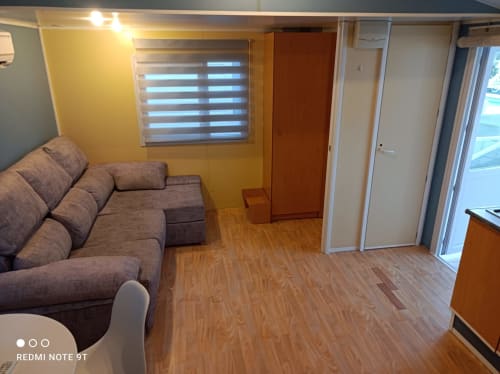 Living room with gray sofa, yellow wall, striped window blinds, wooden floor, and garden view at Bungalow Buda Beach,