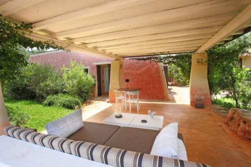 Terrazza ombreggiata presso la Masseria Picca Picca B&B a Morciano di Leuca con divano rigato, edificio rosso e verde circostante