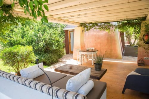 Patio in ombra con sedute a righe, zona pranzo e rigogliosa vegetazione alla Masseria Picca Picca a Morciano di Leuca