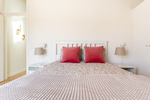 Bright bedroom at Casa Hortas villa in Bordeira with white headboard, red pillows, beige bedding, and matching bedside lamps
