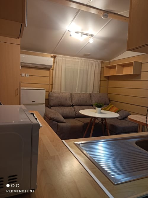 Bungalow D interior at Buda Beach, Chipiona: modern living area with gray sofa, kitchenette, wood paneling, and dining table.