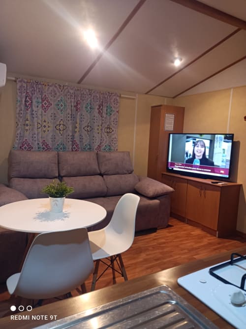 Modern living room in Bungalow E at Buda Beach hotel, Chipiona, with gray sofa, white dining table, flat-screen TV, and