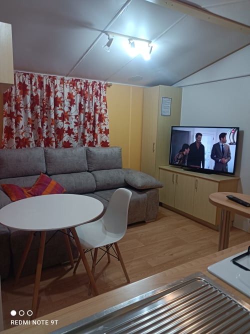 Modern living room with gray sofa, red curtains, flat-screen TV, and dining area in Bungalow F at Buda Beach Chipiona
