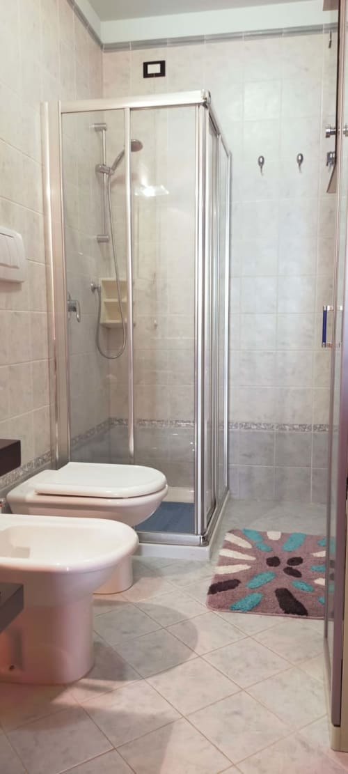 Modern bathroom with glass shower enclosure, white toilet, beige tile walls, and decorative floor mat at Casa Meravigliosa