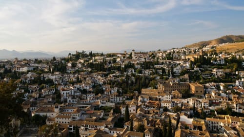 Paisaje urbano de Granada con edificios blancos y terracota descendiendo por la ladera, cipreses y montañas al fondo