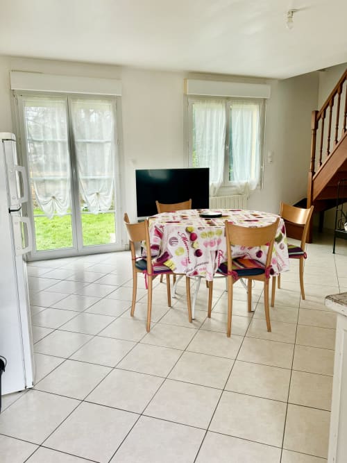 Bright dining area in Le Clos 2 cottage with floral tablecloth, wooden chairs, TV, and garden views through glass doors
