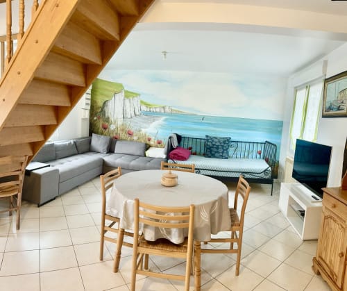 Open-plan living area in Le Clos cottage with coastal mural, gray sectional sofa, round dining table, wooden staircase, and