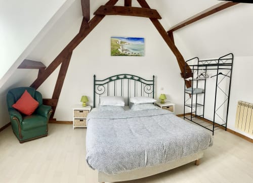 Spacious attic bedroom at Le Clos De La Vallée cottage with exposed wooden beams, iron bed, green armchair, and garden views.