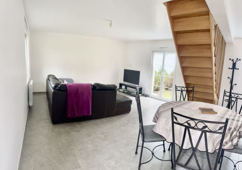 Open-plan cottage living area with black sofa, TV, dining table, and wooden staircase at Le Clos De La Vallée, Bellengreville