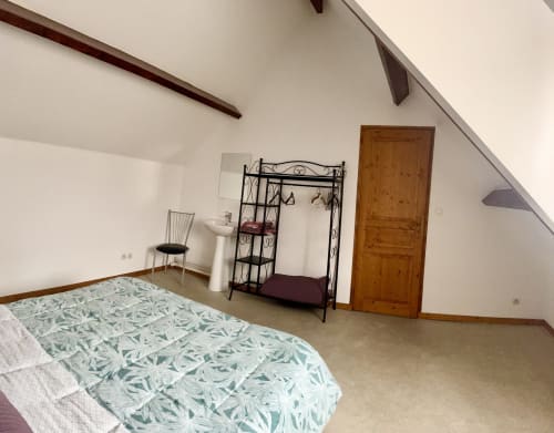 Spacious attic bedroom at Le Clos De La Vallée cottage with bed, metal storage rack, wooden door, and sloped ceiling.