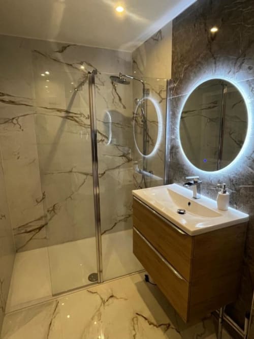 Salle de bain avec du carrelage effet marbre au sol et au mur. Douche à l'italienne et miroir rond lumineux au mur.