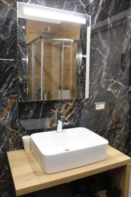 Bagno moderno con lavabo rettangolare bianco su vanity in legno, specchio illuminato e pareti in marmo scuro con venature dorate