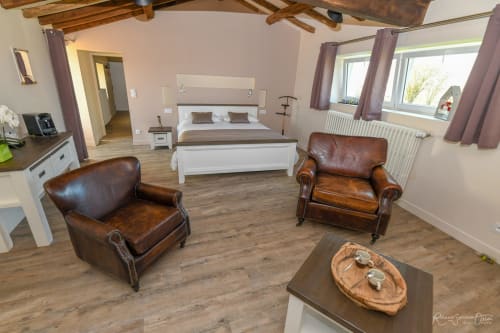 Chambre spacieuse aux Gites De La Grande Borderie avec fauteuils en cuir, lit blanc, poutres en bois et coin détente