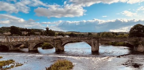 Stone bridge spanning river at Haydon Bridge with rolling hills and rural landscape under blue sky