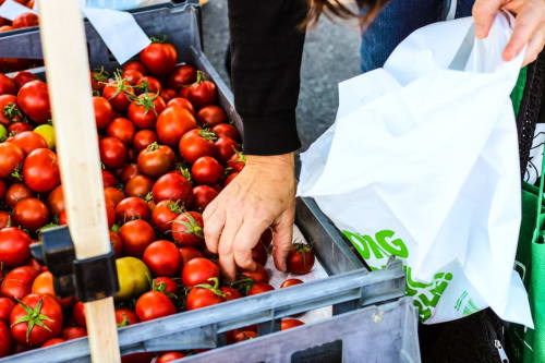 Cliente che sceglie pomodori freschi al mercato di Bari, con busta bianca vicino alla bancarella di frutta e verdura