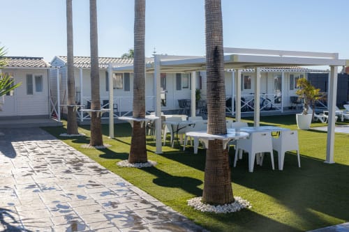 Bungalow Buda Beach garden with white bungalows, palm trees, outdoor dining, and lawn area in Chipiona