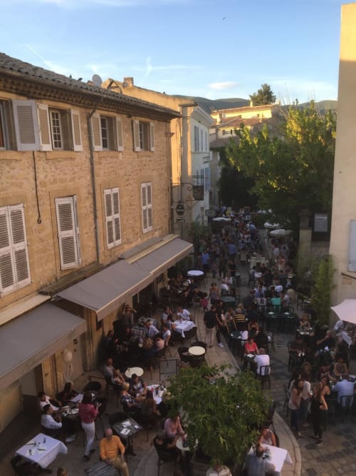 Bustling outdoor cafe scene at AJOUCADOU in Lourmarin, France, with historic stone buildings and diners