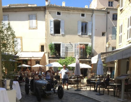 Charming square in Lourmarin with vintage race car, outdoor café seating, white umbrellas, and historic buildings