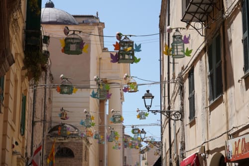 Gabbie colorate appese nella stretta via di Alghero con edifici storici e cupola di chiesa, Catalogna10 Rooms e Loft