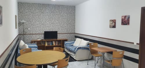 Sala comum do hostel Sunny House em Lagos com camas compartilhadas, mesa redonda, cadeiras de madeira, estante de livros e parede de pedra moderna