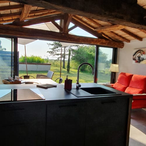 Cuisine de villa moderne avec îlot noir, sièges aux coussins rouges, poutres en bois apparent et grandes baies vitrées surplombant le domaine