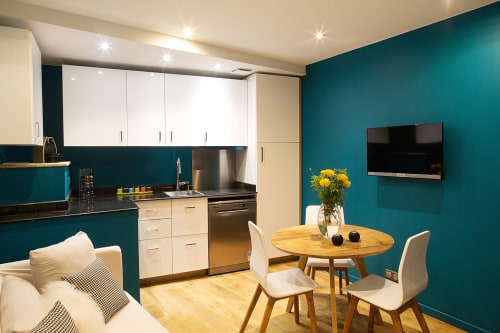 Appartement moderne AJOUCADOU à Lourmarin avec murs turquoise, cuisine blanche, table à manger en bois et télévision murale