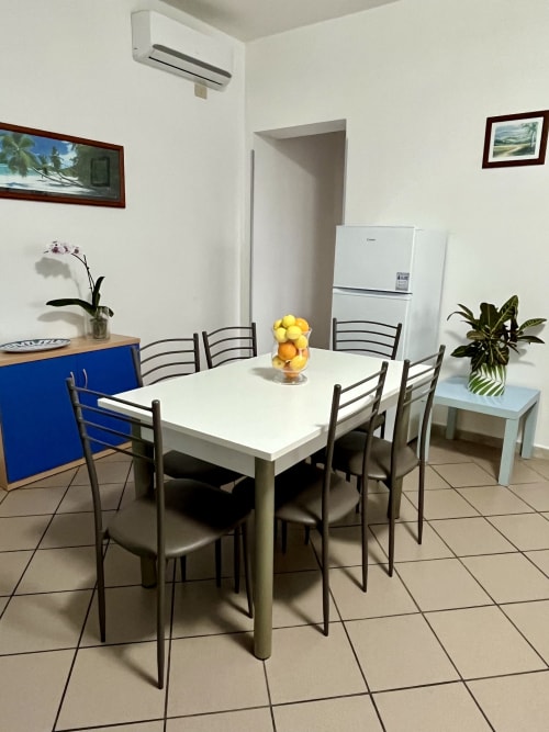Moderna zona pranzo dell'Apartment Tirreno con tavolo bianco, quattro sedie, centrotavola di frutta fresca, piante e arredi bianchi