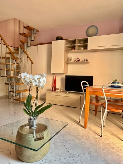 Moderno appartamento su due livelli a Tirrenia con pareti rosa, scala in legno, arredamento crema, TV, orchidee e zona pranzo