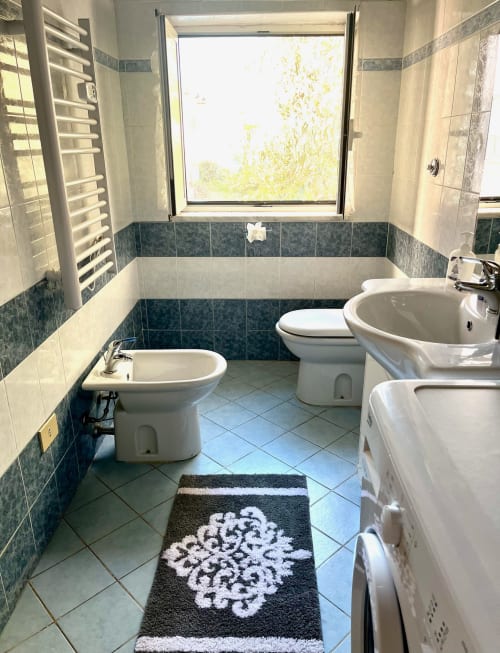 Bagno luminoso presso l'Apartment Tirreno a Tirrenia con pareti piastrellate, tre sanitari bianchi e finestra con vista sul mare