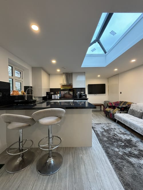 Modern open-plan kitchen and living area with black countertops, cream bar stools, skylight, flat-screen TV, and comfortable