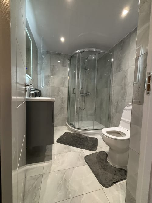 Modern bathroom with curved glass shower enclosure, white toilet, dark bath mats, marble-effect tiles, and illuminated mirror