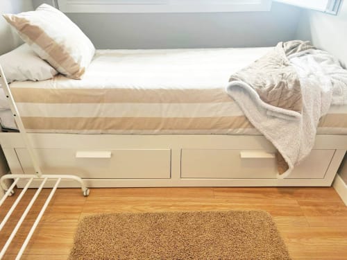 Dormitorio luminoso en apartamento Holibai de Baiona con cama individual, base de almacenamiento beige, ropa de cama blanca y suelo de madera.