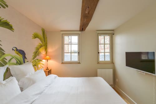 Bright bedroom in Honfleur cottage with white bed, tropical plant mural, wooden beam, TV, and city view windows
