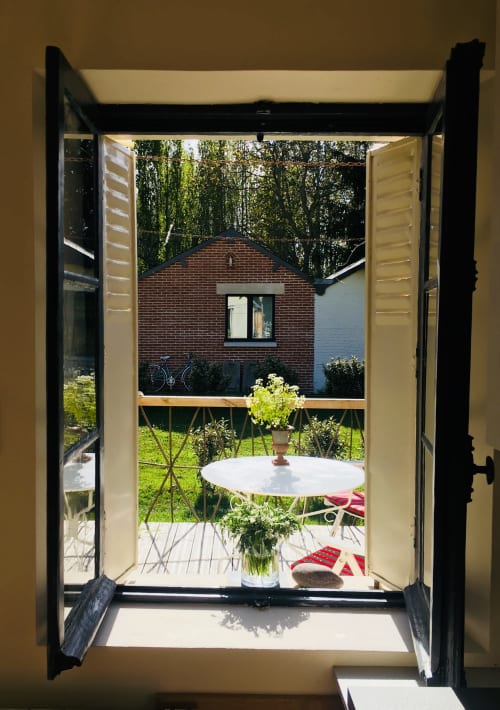 Open doors reveal a lush garden patio at Clairseine cottage with white table, flowers, and brick house beyond