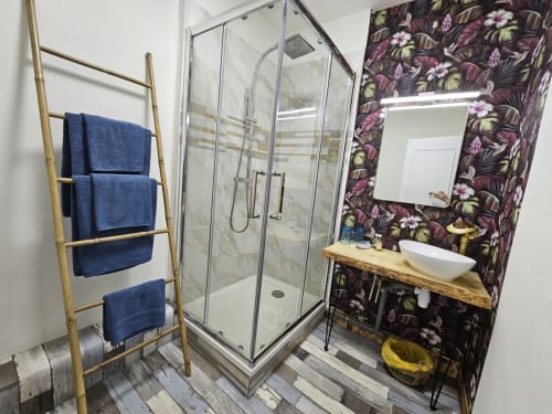 Bagno moderno con carta da parati floreale, doccia con vetrata, mobile lavabo in legno e scaletta portasciugamani blu presso Bastide du Thoré