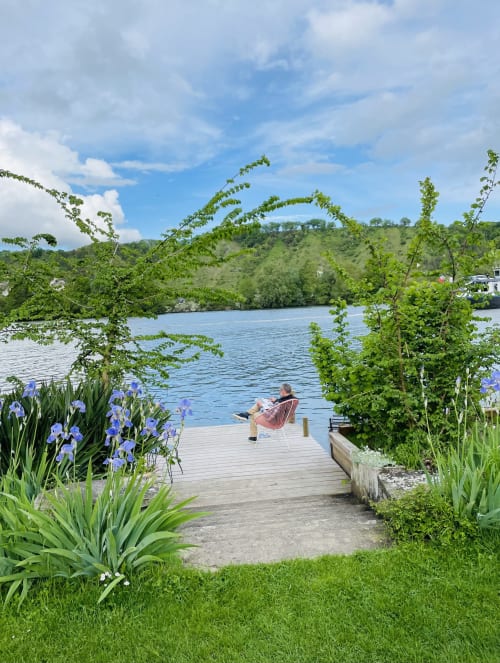 Person relaxing on wooden deck overlooking blue river at Clairseine cottage, Tournedos-sur-Seine, surrounded by lush greenery