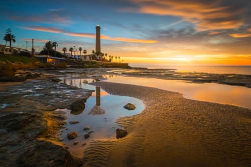 Chipiona lighthouse at golden sunset with tidal pools and palm trees reflecting in sandy beach