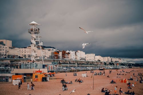 Beachfront promenade with lighthouse, sandy beach, visitors, and seagulls flying overhead under cloudy sky