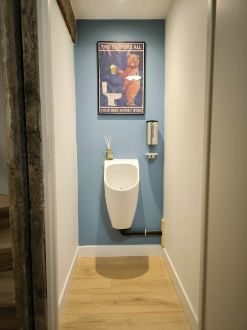 Salle de bains du Repaire Des Darous avec affiche humoristique de bière au-dessus d'un urinoir blanc sur mur d'accent bleu