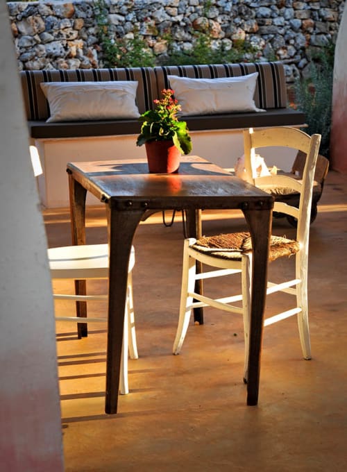 Soleggiata terrazza esterna di Masseria Picca Picca con tavolo scuro, sedie in paglia bianca e muro in pietra vicino a Morciano di