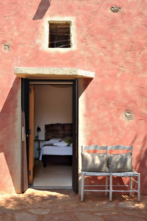 Esterno rosa corallo della camera Gallina a Masseria Picca Picca con porta in legno aperta, sedie bianche e piccola finestra a Morciano