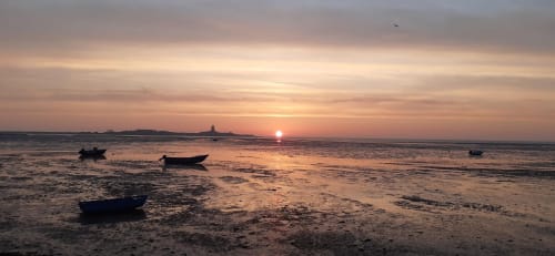 Coucher de soleil sur la plage de Saint-Vaast-la-Hougue avec bateaux amarrés et silhouette du phare à L'Ermitage